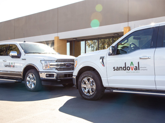 two white trucks parked next to a building, each labeled with Sandoval Elevator's name and logo